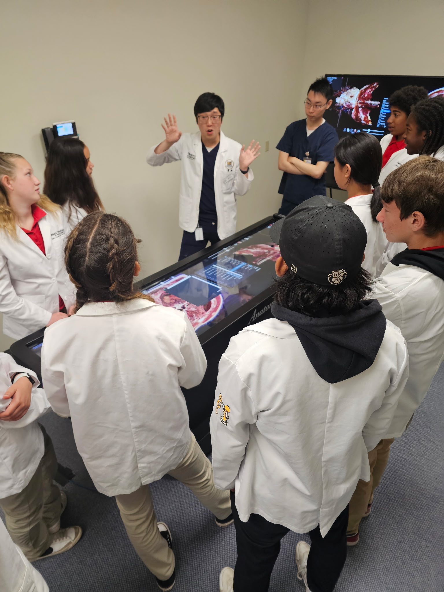 A group of students in white lab coats gather around a digital anatomy table, listening to a person explaining medical details.