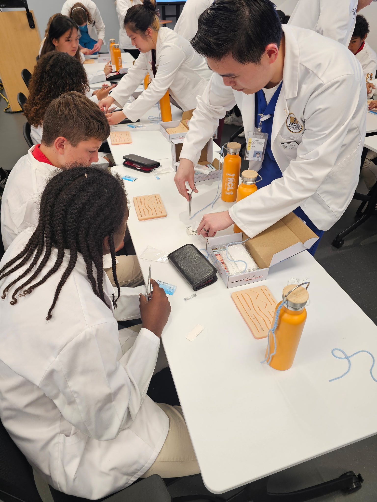 Students in lab coats practice suturing techniques with orange practice pads and tools at a table, guided by an instructor.