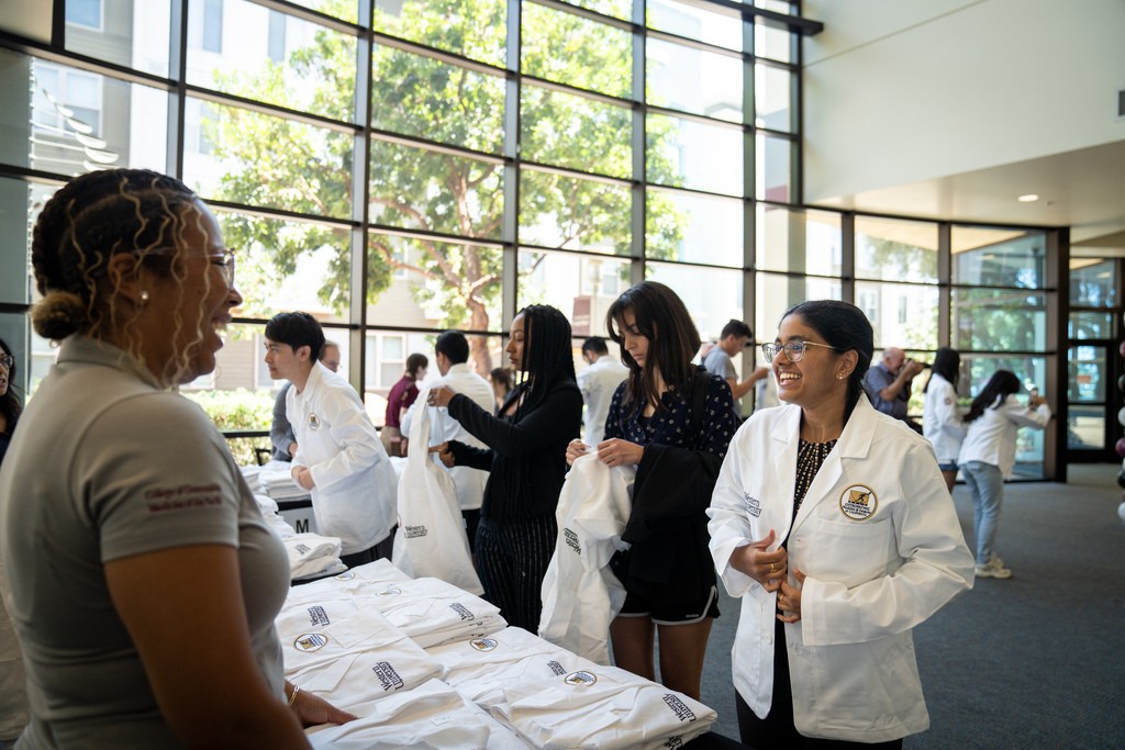 A group of people, some in white coats, are gathered in a well-lit room with large windows. They appear to be engaging in a conversation or event.