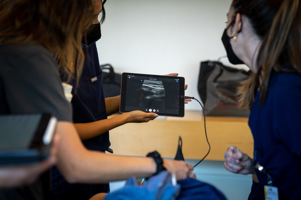 Three people in medical uniforms, wearing face masks, examine an ultrasound image displayed on a tablet device in a clinical setting.