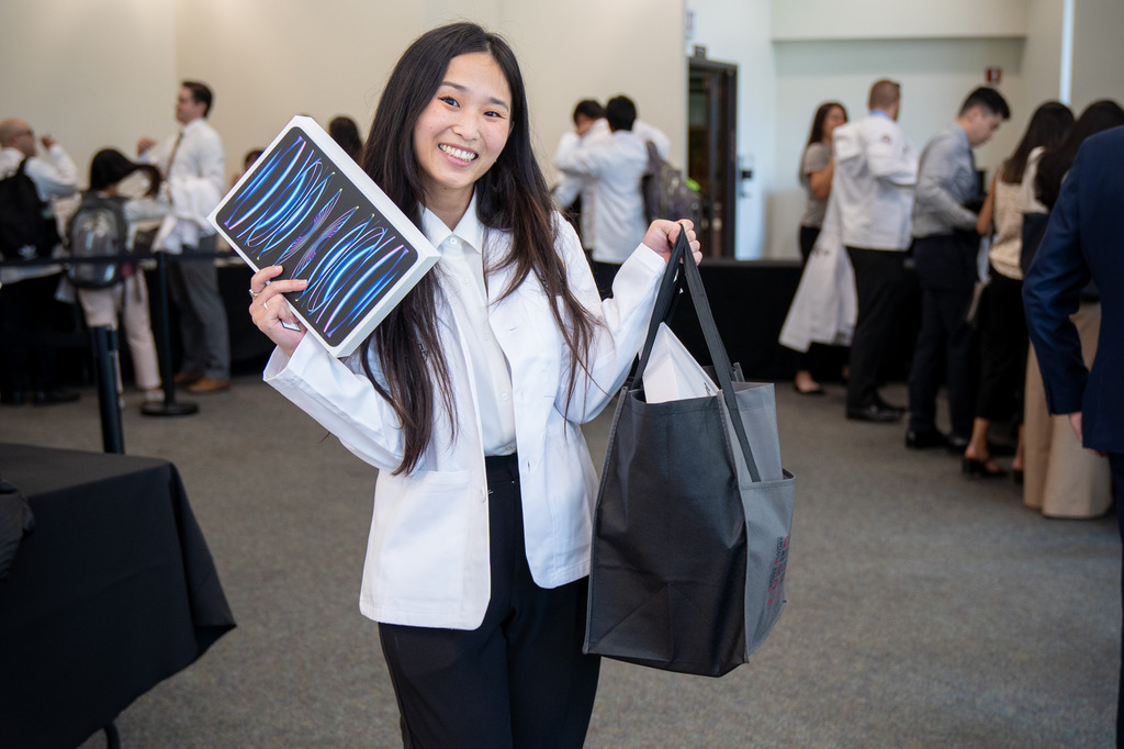 A person in a white coat smiles while holding a tablet and a black bag in a room with other people dressed in white coats.