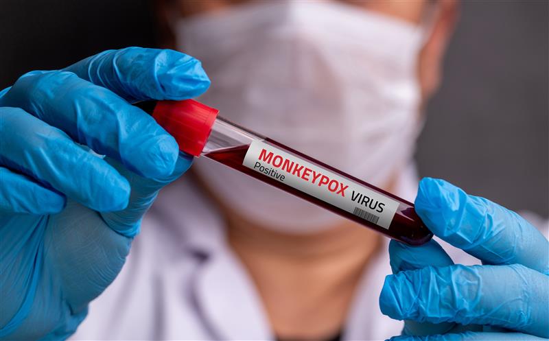 A person wearing a mask and blue gloves holds a test tube labeled 
