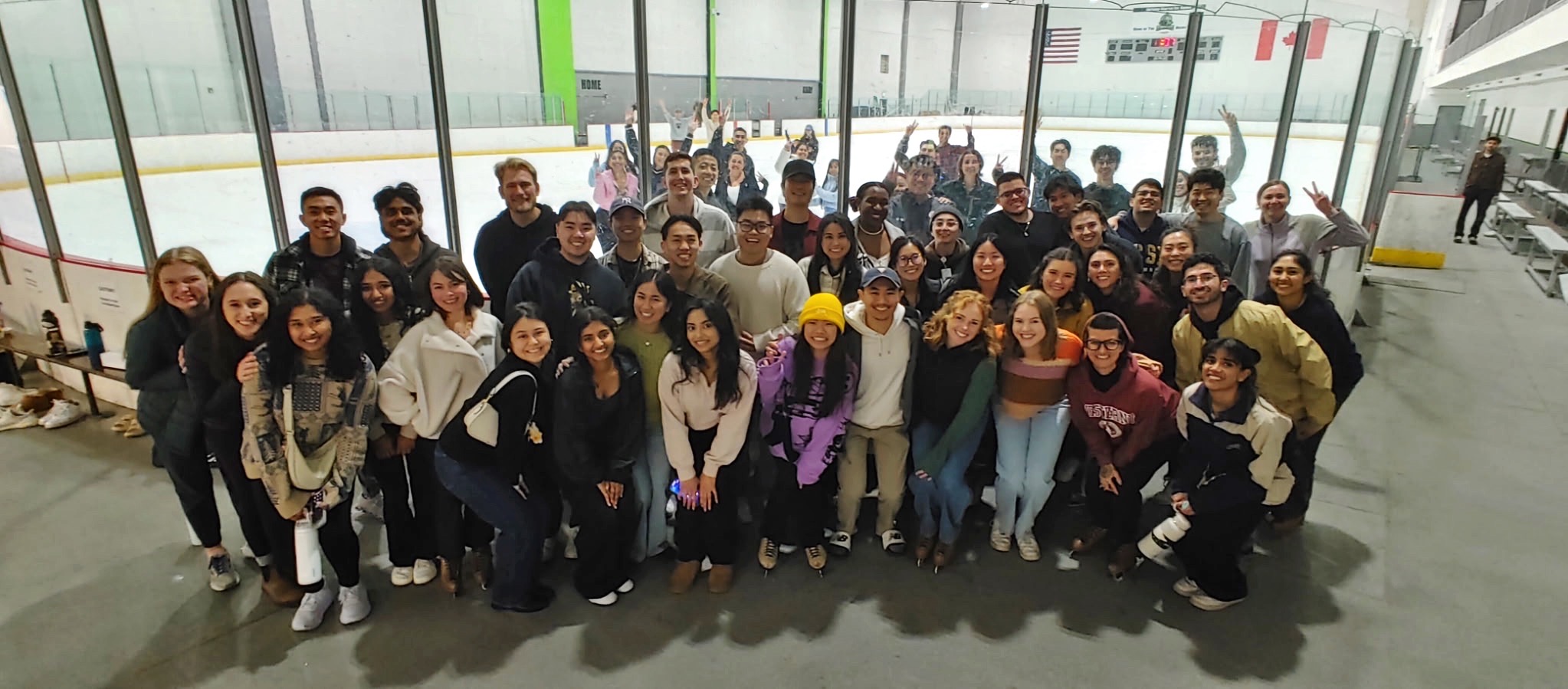 A large group of young adults pose together at an indoor ice rink, smiling at the camera with the ice and rink boards visible behind them.